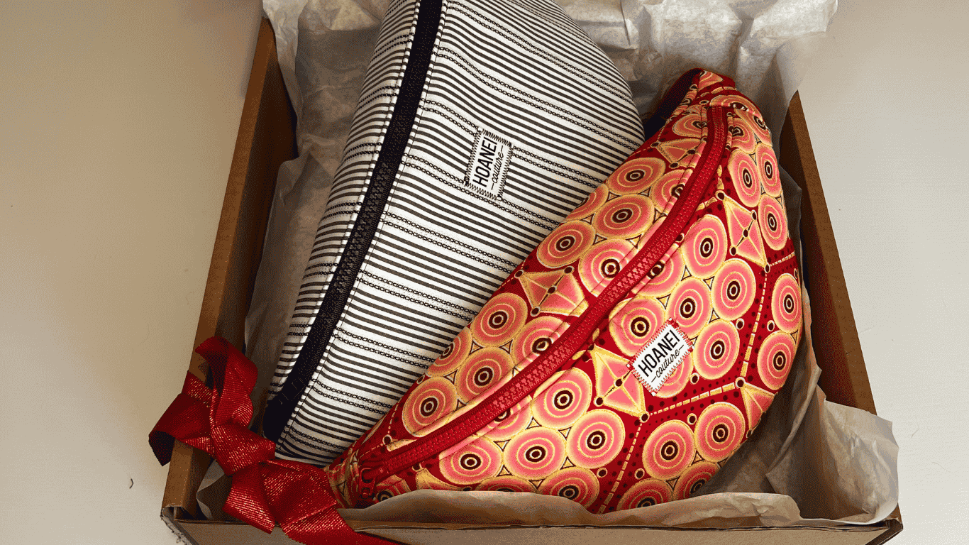 Un colis avec deux sacs bananes artisanales de la marque Hoanei Couture. Un sac est rayé bleu et blanc et l'autre sac est fabriqué à partir de wax rouge et jaune.
